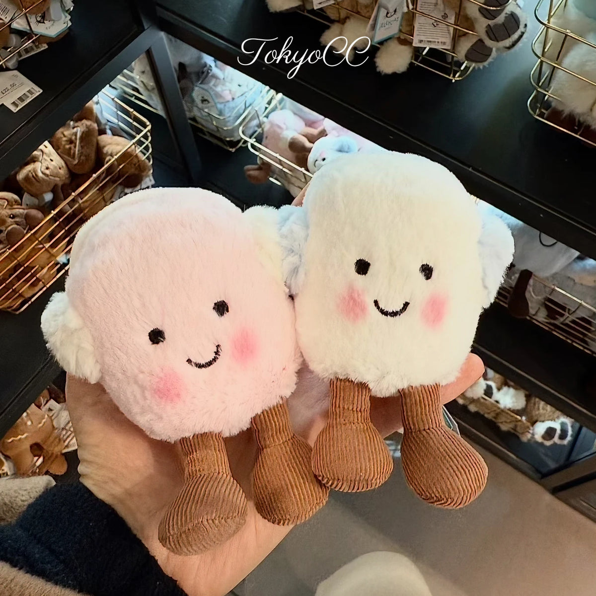 A pair of fluffy pink and white marshmallow plush toys with stitched faces are held in a person's hand.