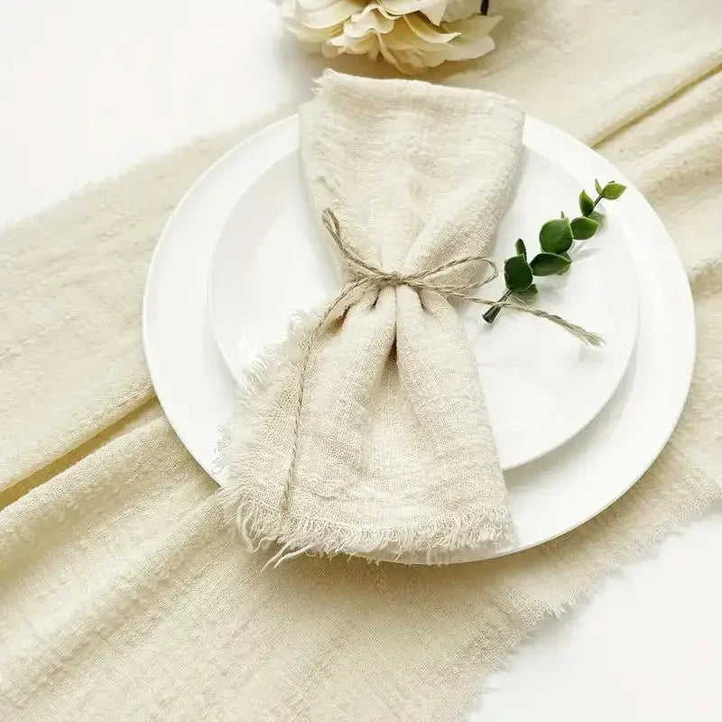 A bundle of handcrafted fringed cotton napkins in a natural beige color resting on a white plate and linen fabric.