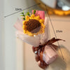 A handmade crochet flower bouquet featuring a large yellow sunflower with a brown center, presented in pink tissue paper.