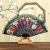 A Spanish style folding lace trim handheld fan with a dark fabric and floral pattern displayed against a light background.