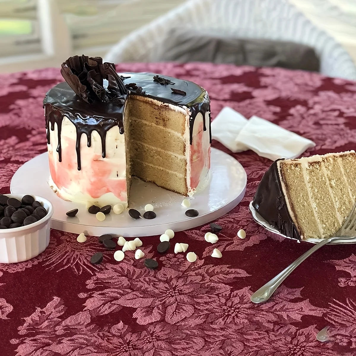 A slice of pink and white cake with chocolate drizzle is served on a plate surrounded by chocolate chips.
