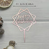 A pink acrylic birthday sign with a geometric shape and text on a wooden surface surrounded by pinecones.