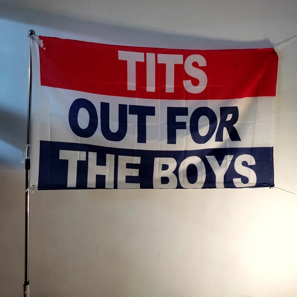 A red and blue flag hangs on a wall with white text that reads TITS OUT FOR THE BOYS
