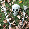 A realistic white skeleton hand lawn stake is shown lying on green grass amidst fallen autumn leaves.
