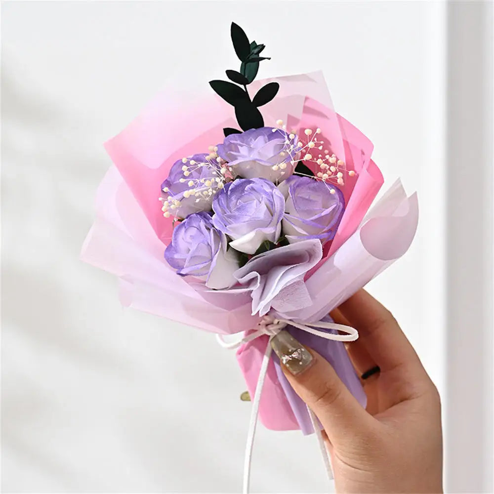 A hand holds a bouquet of handcrafted soap roses wrapped in pink and purple tissue paper.