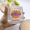 A hand holds a clear glass tumbler with a new achievement sticker on it, next to wooden coasters.