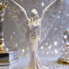 A white figurine of a praying angel with large wings is displayed on a sparkling surface.