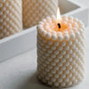 A white candle with a lit wick is set in a cylindrical holder, surrounded by small white bubbles.