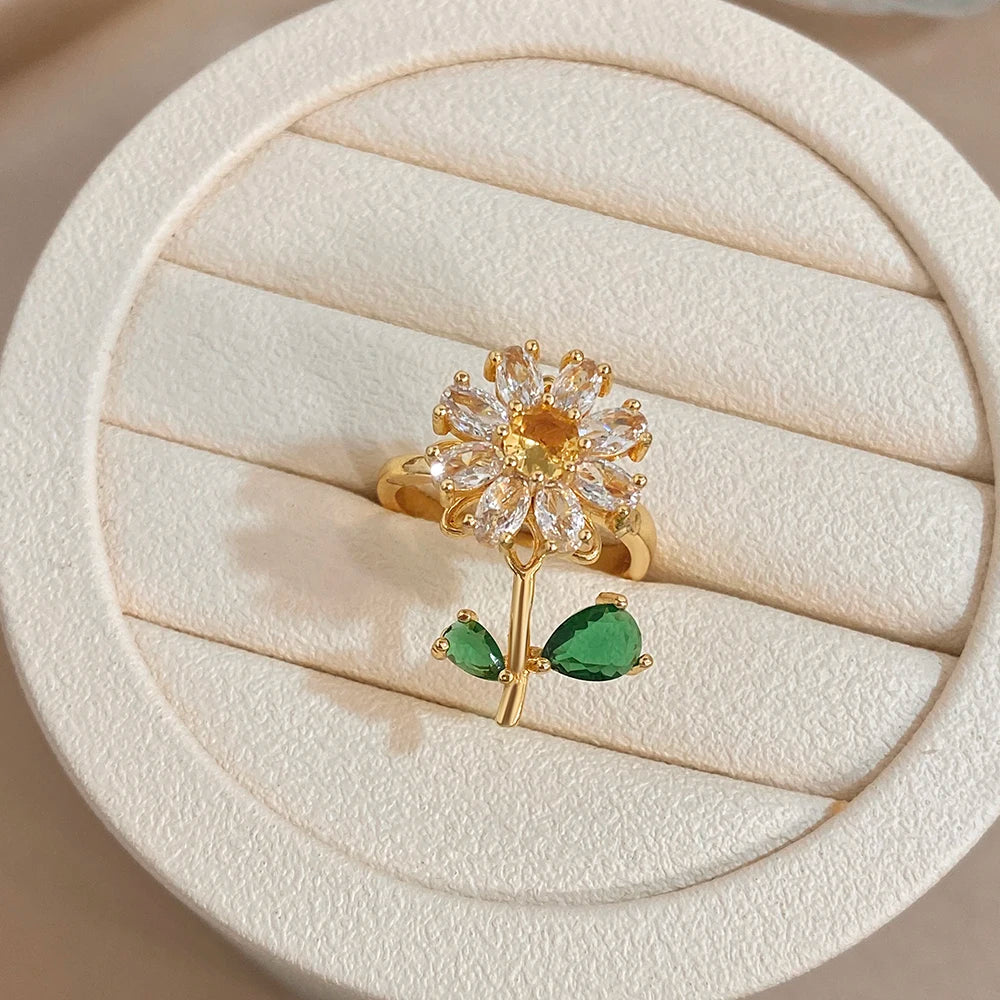 A gold cocktail ring featuring a floral design with clear stones and a green leaf accent, displayed on a light background.