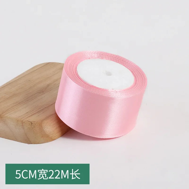 A roll of pink silk satin ribbon with a white center core is shown next to a wooden block.