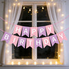 A pink and purple birthday banner strung across a window with string lights and sheer curtains.