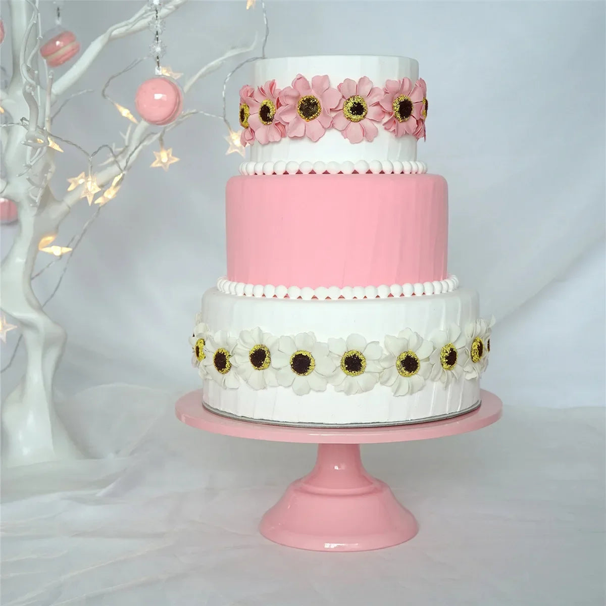 A three-tiered pink and white cake decorated with floral accents displayed on a pink metal stand.