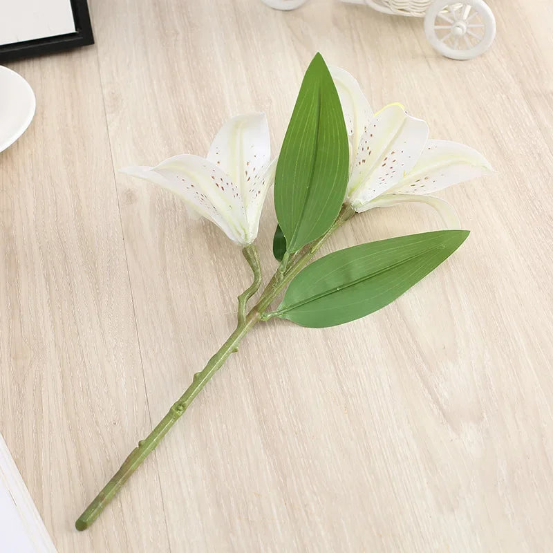 A realistic silk lily flower stem with three white blooms and green leaves on a light wooden surface.