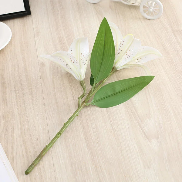 A realistic silk lily flower stem with three white blooms and green leaves on a light wooden surface.