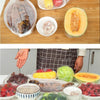 Various fresh vegetables and meats are displayed in reusable plastic food wrap covers on a wooden table.