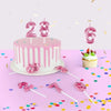 A pink birthday cake decorated with pink dripping frosting and candles, surrounded by pink confetti and decorative candles.