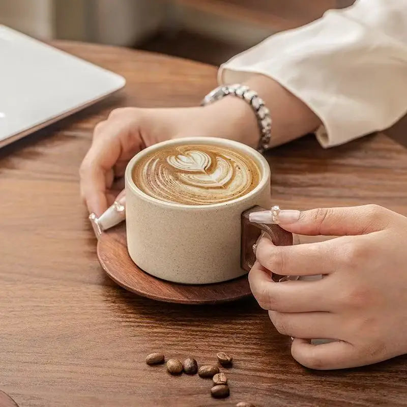 A hand holds a latte in a ceramic cup and saucer on a wooden table.