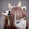 A light brown wig features a headband with cow ears made of white and black faux fur, adorned with gold bells and a ribbon bo