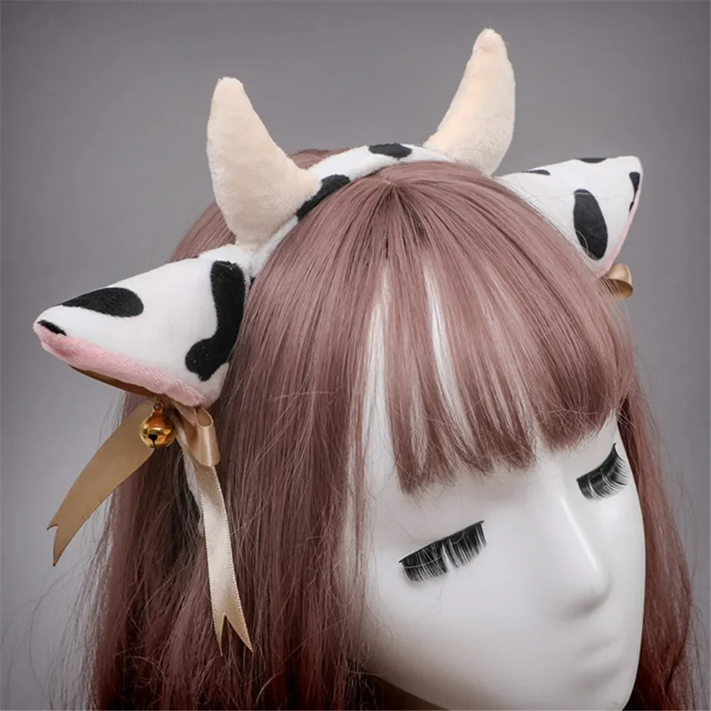 A light brown wig features a headband with cow ears made of white and black faux fur, adorned with gold bells and a ribbon bo
