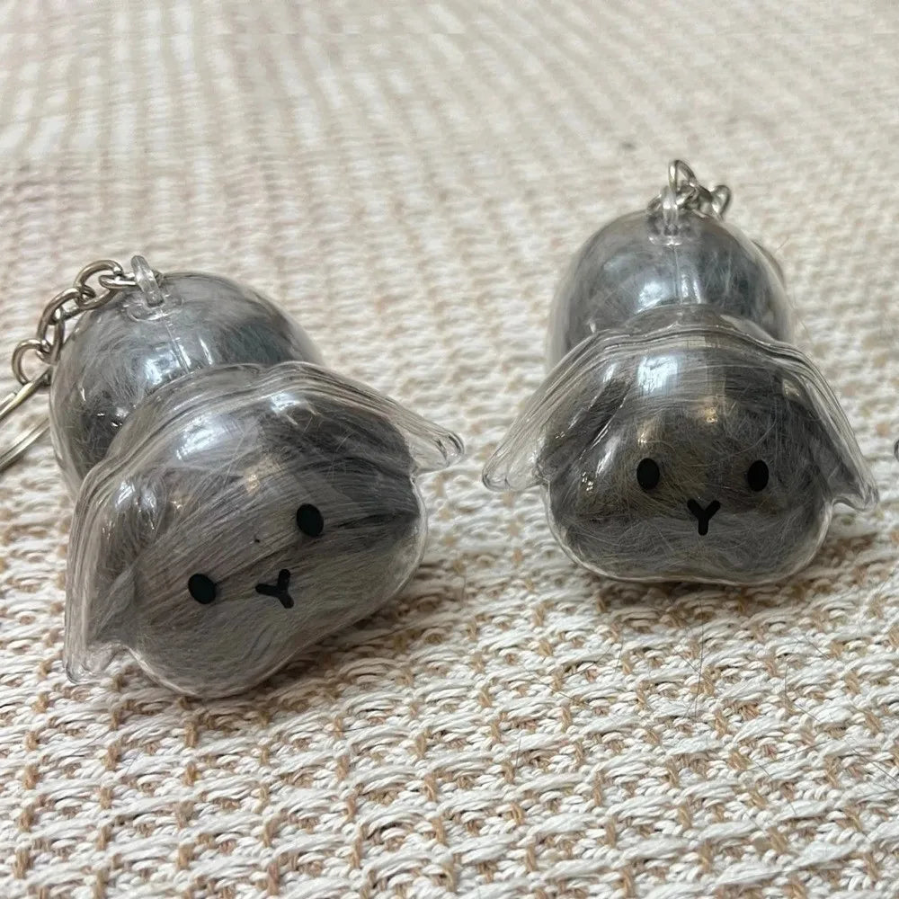 A pair of silver-toned rabbit hair keepsakes or keychains resting on a beige woven fabric surface.