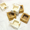 Several small kraft paper favor boxes with natural twine bows are displayed on white fabric.