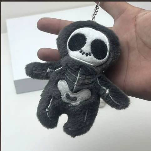A dark gray mummy skeleton plush keychain is held in a person's hand against a white background.