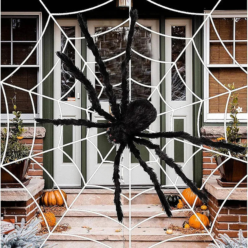 A giant plush black spider decoration is displayed in a white web on a porch with autumn pumpkins.