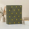 A dark olive green photo album with gold foil heart designs rests on a white surface next to a vase and stacked books.