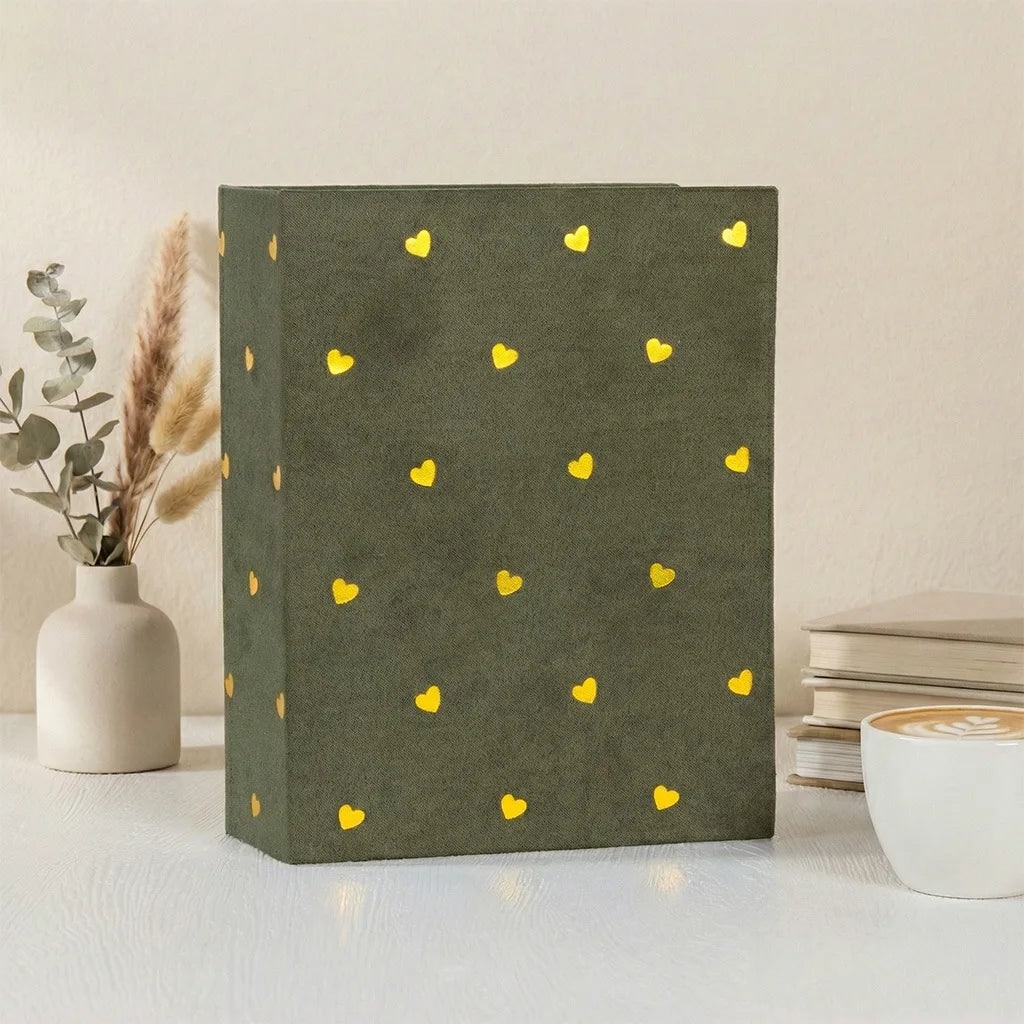 A dark olive green photo album with gold foil heart designs rests on a white surface next to a vase and stacked books.