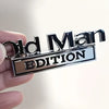 A hand holds a silver metal emblem with the text Old Man Edition on a rectangular plaque.