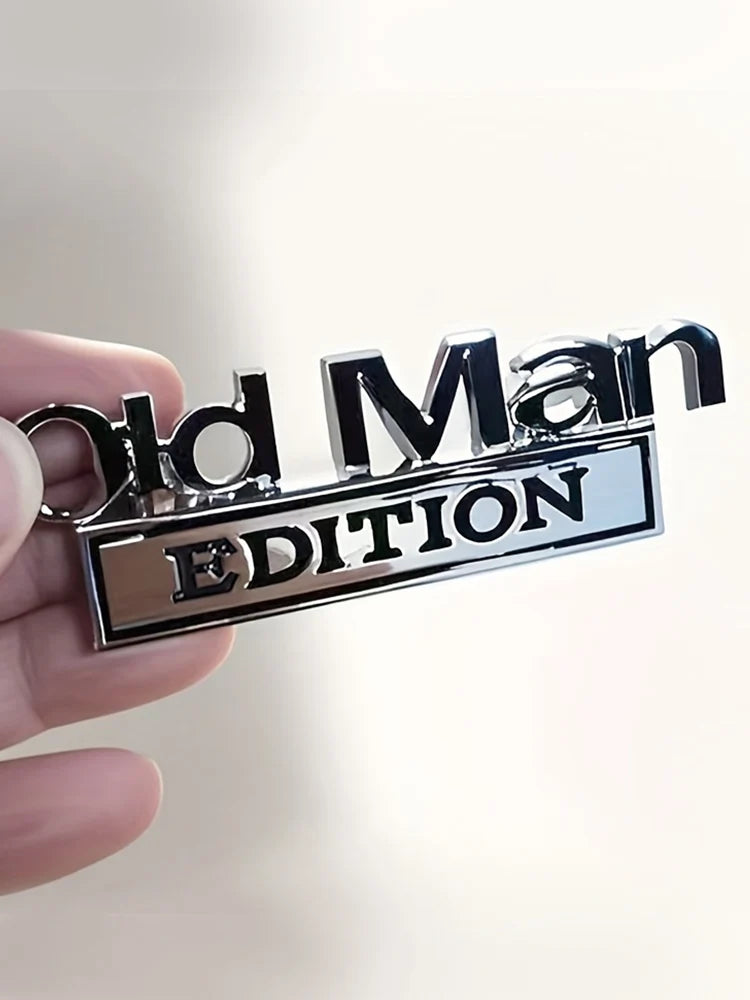 A hand holds a silver metal emblem with the text Old Man Edition on a rectangular plaque.