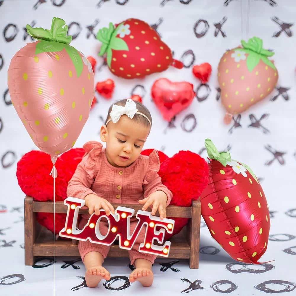A baby sits in front of a display of pink and red strawberry foil balloons with heart accents.