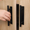 A hand is pulling open a light wood cabinet door using a black, no-drill adhesive handle.