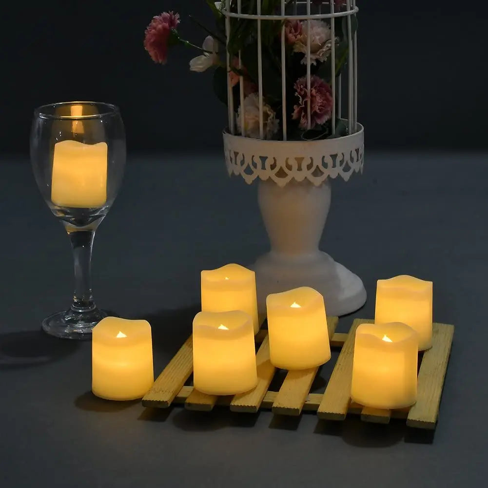 A set of twenty-four glowing yellow LED tea light candles arranged on a wooden tray next to a glass and a floral arrangement.