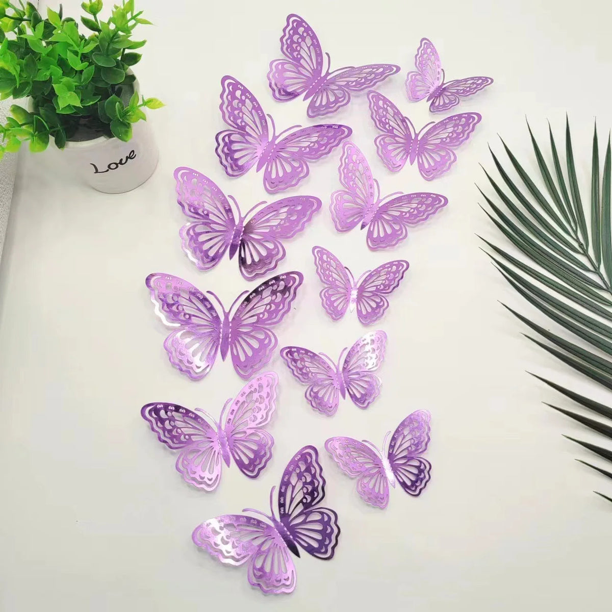 A set of twelve purple and lilac 3D butterfly wall stickers scattered on a white surface next to a green plant.