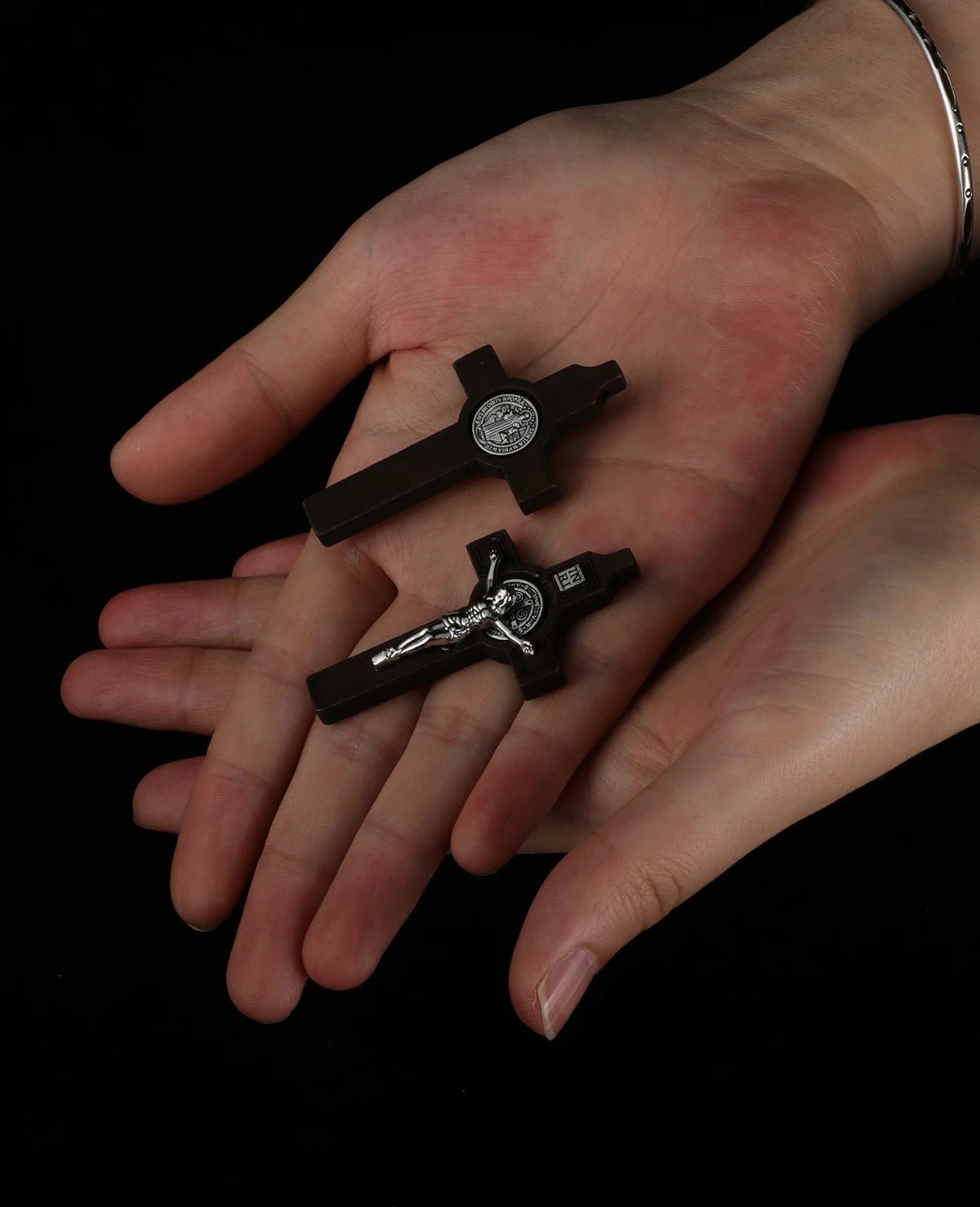 A hand holds a dark wooden St. Benedict crucifix pendant against a black background.