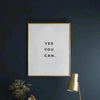 A framed motivational quote print on white canvas with gold frame hanging on a dark blue wall.