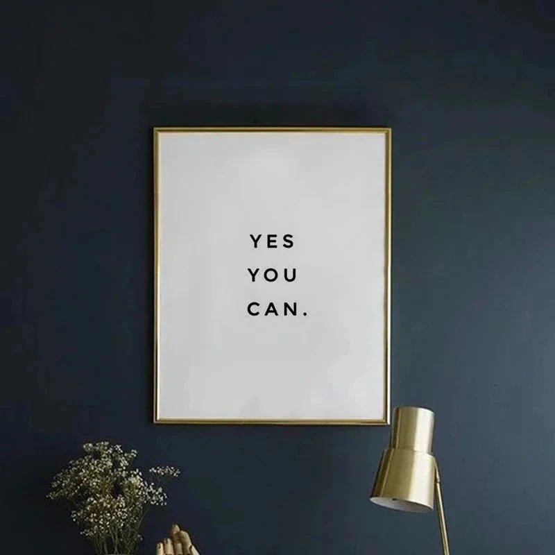 A framed motivational quote print on white canvas with gold frame hanging on a dark blue wall.