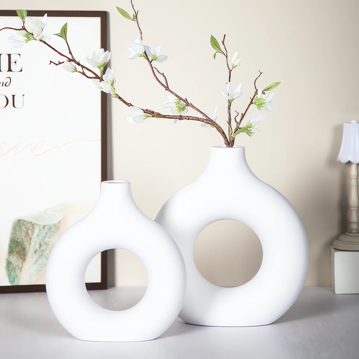 A white ceramic vase with a natural branch of white blossoms is displayed on a light surface.