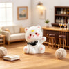 A white resin cat keychain with pink accents sits on a wooden table next to a ball of yarn.