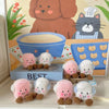 A set of pink, white, and blue marshmallow plush toys displayed in front of a background featuring a brown dog and green plan
