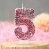 A pink glitter number five candle with a silver crown topper displayed on a wooden surface.