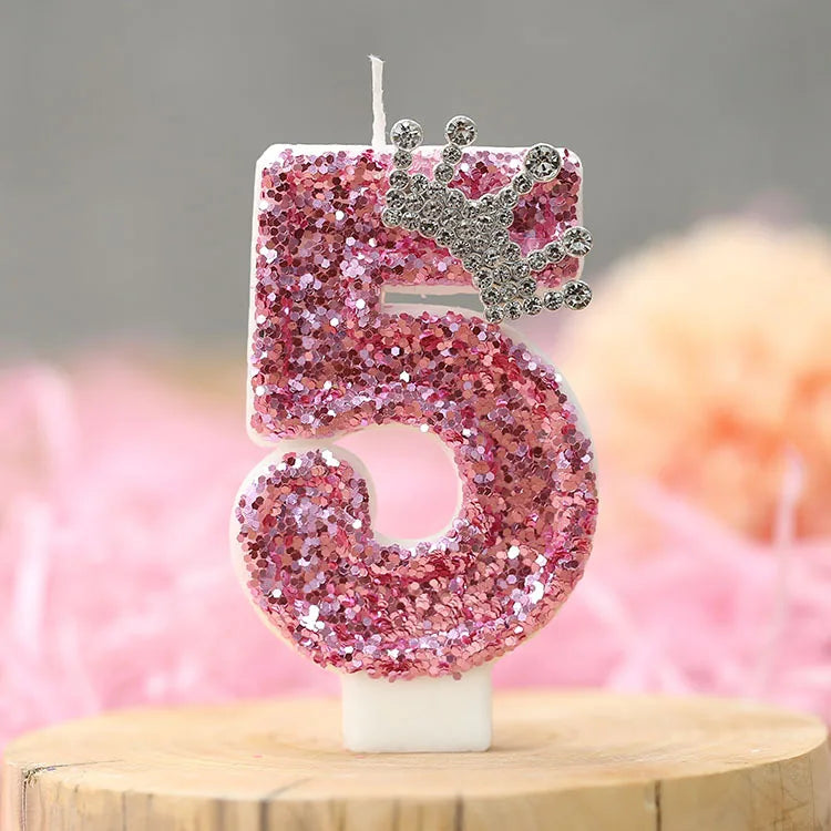 A pink glitter number five candle with a silver crown topper displayed on a wooden surface.