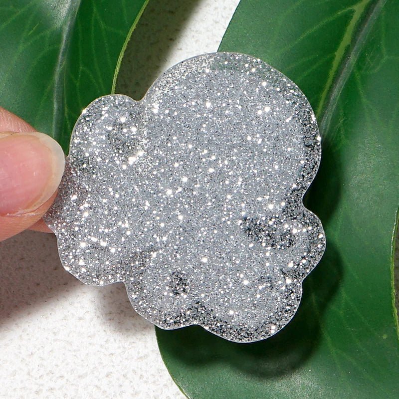 Silver glitter acrylic charm shaped like a cloud being held by a finger against a green leaf background