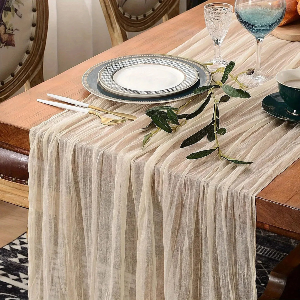 A beige cheesecloth table runner draped over a wooden table set for a meal with plates and glassware.