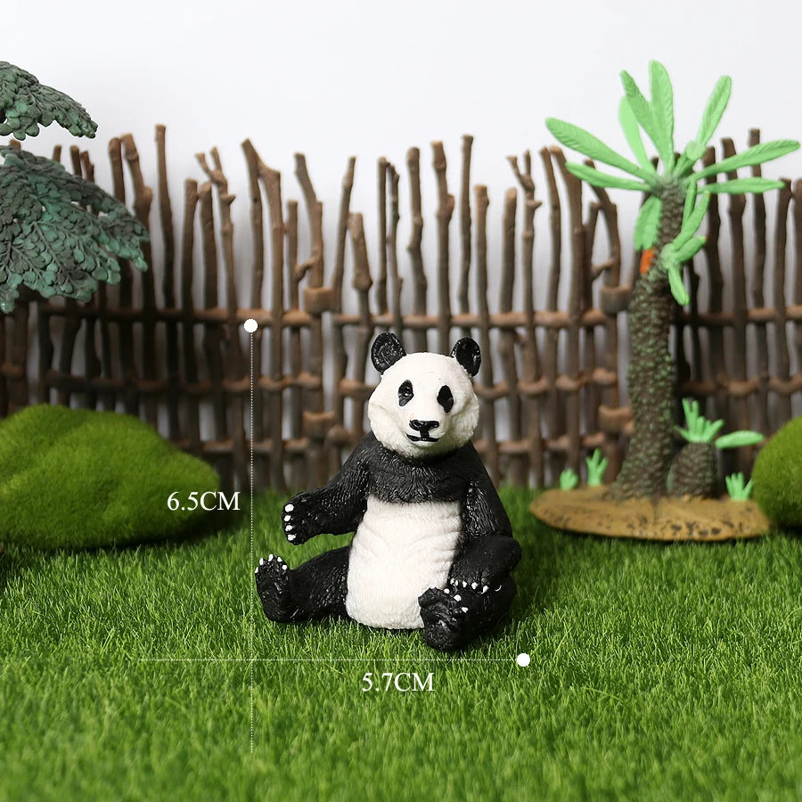 A realistic panda figurine sitting on green grass in front of a wooden fence and palm tree.