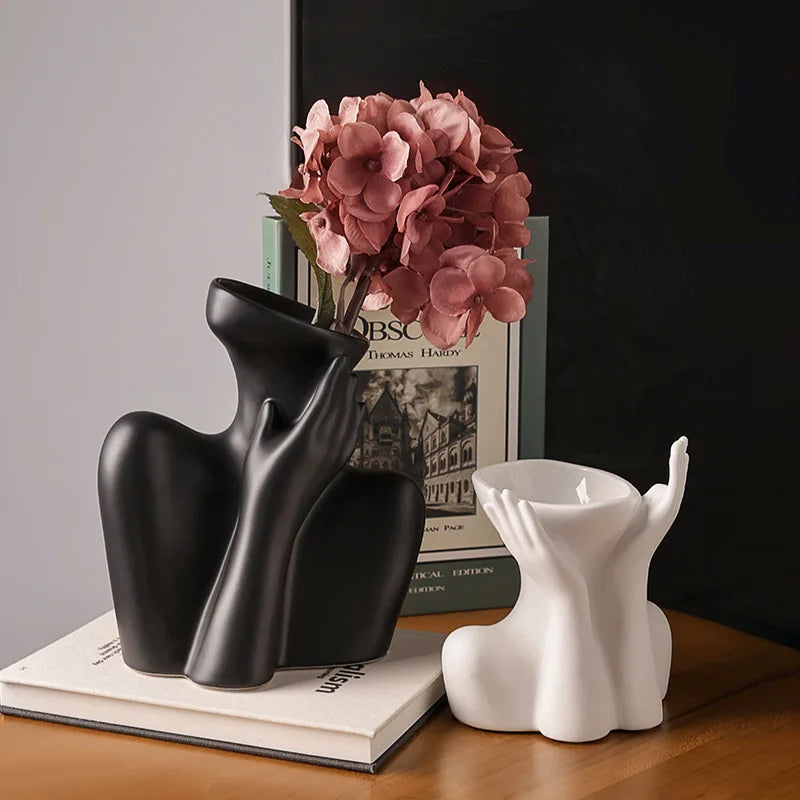 A black ceramic figural vase shaped like an artistic human form sits next to a white candle holder on a wooden surface.