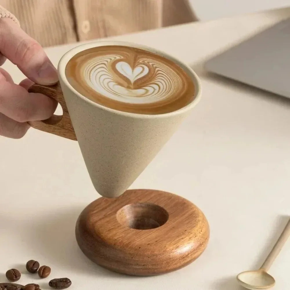 A hand holds a ceramic coffee mug with latte art on top, resting above a wooden coaster.
