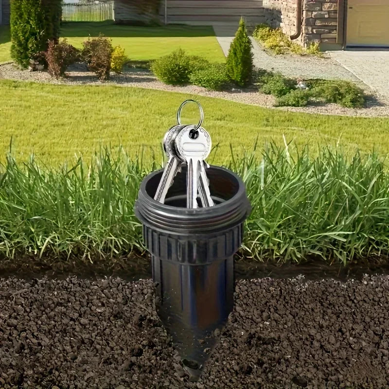 A silver key hider with a handle is placed in a dark pot in a grassy garden area.