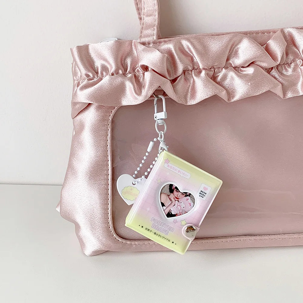 A pink satin pouch holds a clear photo album keychain with a heart design.